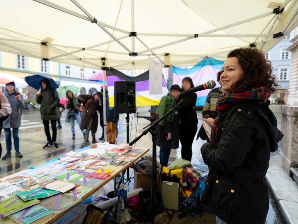 Kathi Bacher steht unter einem Pavillon hinter einem Tisch mit Flyern vor einem Mikrofon und lächelt. Im Hintergrund hängen eine trans und eine nonbinary Flagge. Es stehen einige Menschen unter dem Pavillon oder davor und schauen zu Kathi.